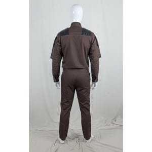 May include: A brown jumpsuit with black shoulder accents and decorative stitching on the sleeves. The outfit has a high collar and long sleeves. The pants are straight-legged.