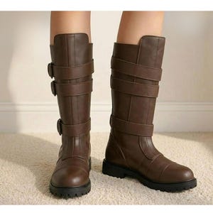 Peut inclure: Une paire de bottes mi-mollet marron dot&eacute;es de trois brides horizontales avec boucles assorties sur la tige. Les bottes pr&eacute;sentent un bout arrondi, une semelle noire plate et une finition lisse, montant jusqu'au milieu du mollet.