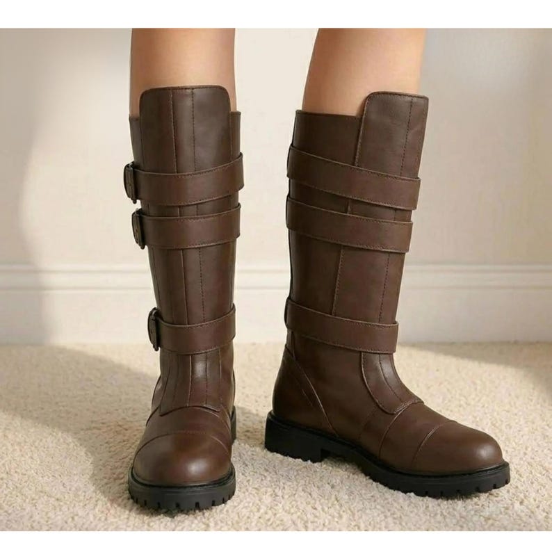 Peut inclure: Une paire de bottes mi-mollet marron dot&eacute;es de trois brides horizontales avec boucles assorties sur la tige. Les bottes pr&eacute;sentent un bout arrondi, une semelle noire plate et une finition lisse, montant jusqu'au milieu du mollet.