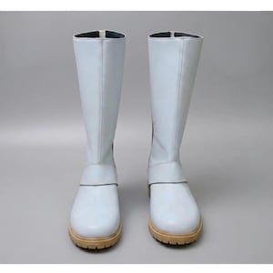 May include: A pair of light blue knee-high boots. The boots have a smooth, leather-like texture, a strap detail around the ankle, and a tan sole. The boots are displayed against a plain gray background.