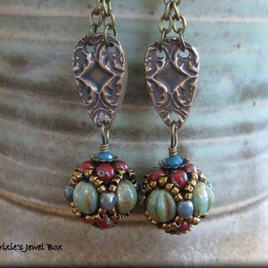May include: A pair of dangle earrings with a bronze-colored, ornate, teardrop-shaped charm and a round, beaded charm with turquoise, red, and gold beads.