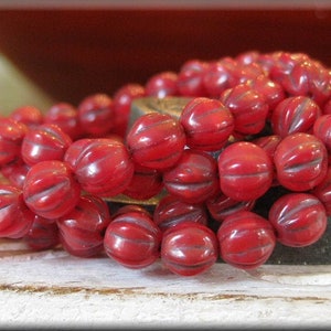 May include: A strand of red glass beads with a ribbed texture. The beads are round and have a slight shine to them.