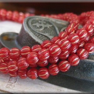 May include: A strand of red glass beads with gold accents. The beads are round and have a ribbed texture. The beads are strung on a wire and are lying on a dark surface.