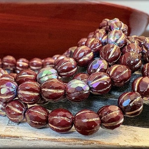 May include: A strand of maroon glass beads with gold accents. The beads are faceted and have a ribbed texture. The beads are strung on a clear thread.