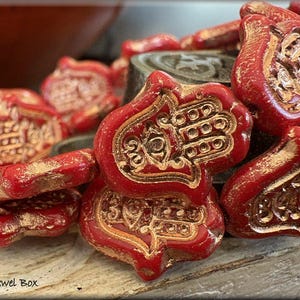 May include: A string of red glass beads with gold accents. Each bead is shaped like a hamsa hand with an eye in the centre. The beads are arranged in a pile on a wooden surface.