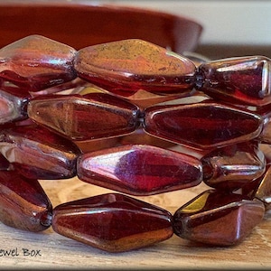 May include: A multi-strand bracelet featuring faceted, reddish-brown beads. The beads have an iridescent sheen, reflecting hues of purple and gold. The bracelet is made of several rows of beads, creating a bold, eye-catching accessory. The text "Trixie's Jewel Box" is in the bottom left corner.