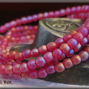 May include: A close-up of a strand of pink iridescent glass beads. The beads are small and round, and they have a subtle shimmer to them. The beads are arranged in a circular pattern, and they are lying on a dark surface.