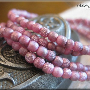 May include: A close-up of a strand of pink iridescent glass beads. The beads are round and have a textured surface. The beads are arranged in a circular pattern and are lying on a textured surface.