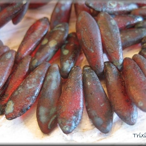 May include: A close-up of a strand of red and blue glass beads. The beads are shaped like long, narrow teardrops and have a mottled, textured surface.