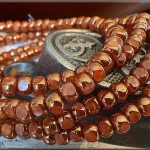 May include: Strands of copper-colored beads are artfully arranged, showcasing their glossy, faceted surfaces. The beads are strung together, creating a layered effect. The image is of a jewelry item, possibly a necklace or bracelet.