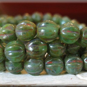May include: A strand of green glass beads with a striated pattern. The beads are round and have a slightly iridescent finish.