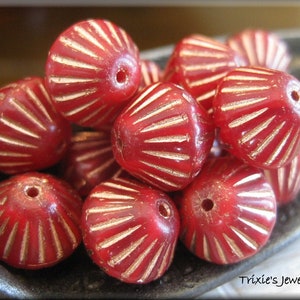 May include: A collection of red beads with a radial design and gold-colored accents. Each bead has a hole for stringing. The beads are in a small, dark-colored dish. The text "Trixie's Jewel Box" is visible in the bottom right corner.