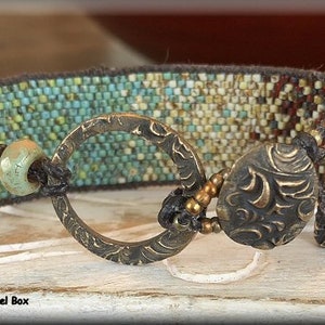 May include: A beaded bracelet with a turquoise and brown color scheme. The bracelet features a large, ornate, circular clasp and a smaller, ornate, teardrop-shaped charm.