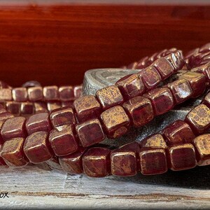 May include: Strands of square beads in a deep burgundy color with a metallic gold finish. The beads are arranged in a spiral pattern, showcasing their textured surfaces. The image includes the text "Trixie's Jewel Box".