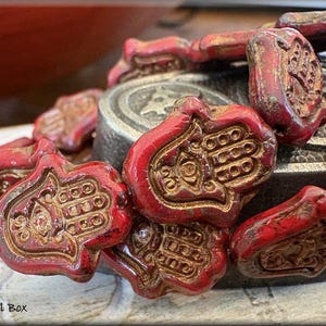 May include: A close-up of a red and gold hamsa hand charm necklace. The hamsa hand is a symbol of protection and good luck. The necklace is made of ceramic beads and has a distressed finish.