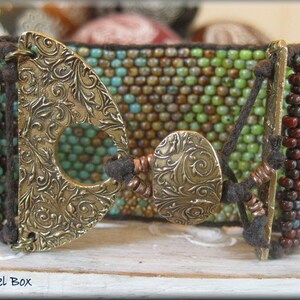 May include: A beaded bracelet with brown, green, and turquoise beads. The bracelet has two large, ornate, gold-toned metal clasps with a floral design.