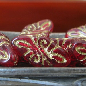 May include: Five red glass beads with gold accents and intricate designs. The beads are arranged in a row on a grey metal surface.