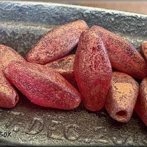 May include: A close-up of a group of red and gold glass beads. The beads are shaped like elongated diamonds and have a textured surface.