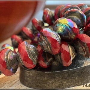 May include: Close-up of a collection of faceted red and iridescent beads. The beads are arranged on a dark, textured surface. The beads have a metallic sheen and are likely part of a jewelry design.