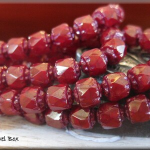 May include: A strand of deep red glass beads with a faceted, square shape. The beads are strung together to create a necklace or bracelet.