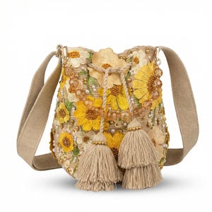 Peut inclure: Sac bandoulière beige avec un motif floral. Le sac présente des tournesols jaunes, des perles blanches et dorées, et deux glands beiges. Le sac a une fermeture à cordon et une bandoulière beige.