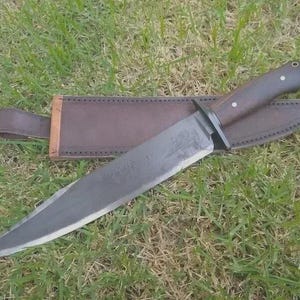 May include: A large, handmade knife with a dark wooden handle and a wide, gray blade. The knife rests in a brown leather sheath with a belt loop. The knife and sheath are set against a backdrop of green grass.