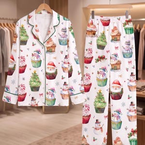 May include: A white pajama set consisting of a long-sleeved button-down shirt with a collar and matching pants. The fabric features a repeating pattern of Christmas-themed cupcakes, including designs of snowmen, Santa Claus, and pine trees.