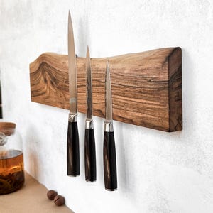 Puede incluir: Un soporte de madera para cuchillos con tres cuchillos. El soporte es de madera oscura y natural con un diseño de borde vivo. Los cuchillos tienen hojas plateadas y mangos marrones oscuros. El soporte está montado en una pared blanca.