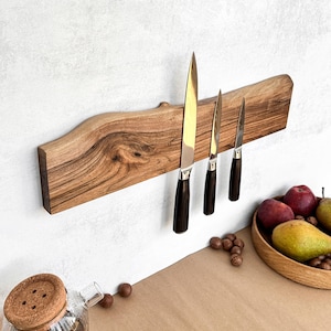 Puede incluir: Un soporte de cuchillos de madera con tres cuchillos. Los cuchillos tienen mangos marrones oscuros y hojas plateadas. El soporte está montado en una pared blanca. Una tetera de vidrio con tapa de corcho está en primer plano.