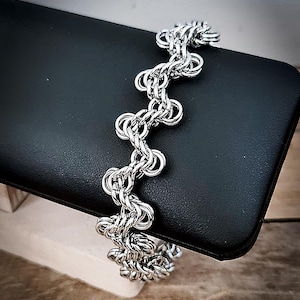 May include: A silver-toned chainmail bracelet featuring a repeating, interlocking circular link pattern. The bracelet is constructed from numerous small, polished metal rings connected in a continuous, flexible, and wavy chain design.