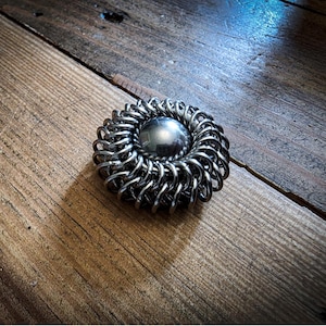 May include: A circular metallic object featuring a smooth, polished dark grey sphere at the centre, surrounded by a dense, intricate border of interlocking silver-toned metal rings that create a textured, chainmail-like pattern around the core.