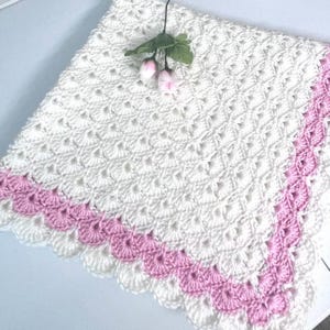 May include: A handmade, white crochet blanket with a scalloped pink border. The blanket features a textured, open weave pattern. A sprig of pink flowers rests on the blanket, adding a delicate touch. This cozy blanket is perfect for a baby or as a decorative throw.