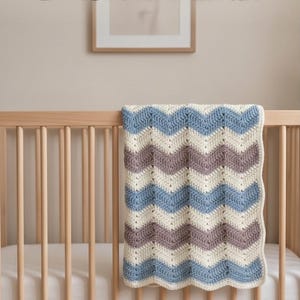 Può includere: Una coperta all'uncinetto con un motivo a chevron ripetuto in strisce orizzontali alternate di color crema, viola polvere e azzurro. La coperta è drappeggiata sulle doghe in legno di una culla, mostrando una lavorazione a maglia testurizzata.