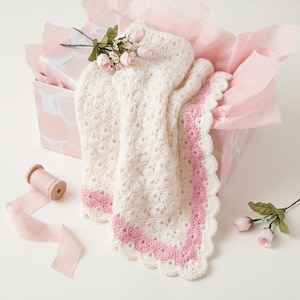 May include: A cream-colored crocheted blanket featuring a shell stitch pattern and a scalloped border in soft pink. The blanket is presented in a gift box with pink tissue paper, accompanied by a spool of pink ribbon and small artificial pink roses.
