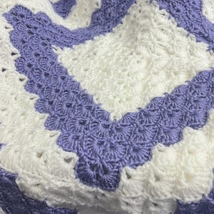 Puede incluir: Una manta de ganchillo hecha a mano con un patrón de chevron en blanco y lavanda. La manta está hecha con hilo suave, mostrando un diseño texturizado. La combinación de colores es una combinación de blanco y morado claro.