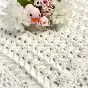 May include: A white crocheted textile with an openwork pattern, topped with a small pink duck figurine, several light pink artificial rosebuds, and two large white artificial chrysanthemum flowers with green stems and leaves.