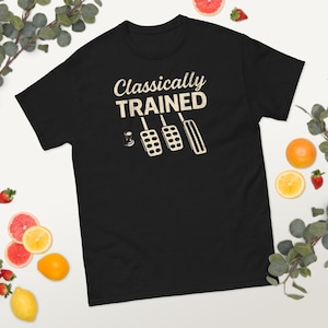 Może przedstawiać: Czarny t-shirt z napisem "Classically TRAINED" w stylu vintage, wraz z ilustracją pedałów samochodowych w kolorze kremowym. Koszulka jest otoczona owocami.