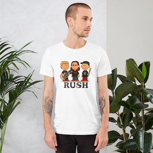 May include: White t-shirt featuring a cartoon illustration of three musicians with the word "RUSH" in black block letters. One character plays a guitar, another wears glasses, and the third wears a cap and holds a drumstick.