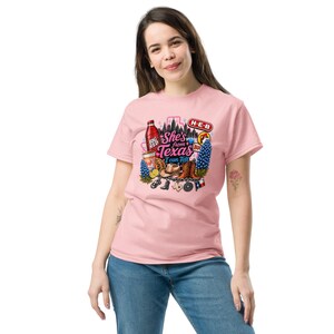 Może przedstawiać: Jasnoróżowy t-shirt z krótkim rękawem z nadrukiem graficznym zawierającym tekst 'She's from Texas I can Tell'. Wzór zawiera ilustracje butelki napoju Big Red, kowbojskiego kapelusza, kowbojek, niebieskich kwiatów oraz różnych symboli stanu Teksas.