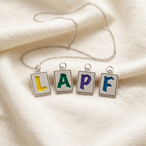 May include: A silver necklace spelling "LAPF" with four square pendants. Each pendant has a different colour: yellow, green, purple, and blue. The necklace is displayed on a cream-coloured fabric.