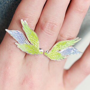 Pu&ograve; includere: Un paio di orecchini a forma di ali argentate. Ogni orecchino presenta un design con dettagli in smalto verde lime e azzurro. Le ali sono esposte su una mano, mettendo in risalto il design e il colore dei gioielli.