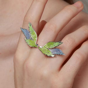 Pu&ograve; includere: Anello in argento con design ad ali stilizzate, con dettagli in smalto verde e blu. Le ali dell'anello avvolgono il dito, creando un gioiello unico e artistico.