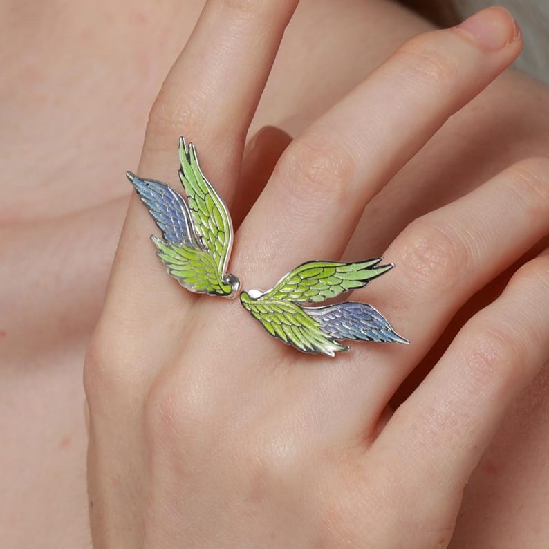 Pu&ograve; includere: Anello in argento con design ad ali stilizzate, con dettagli in smalto verde e blu. Le ali dell'anello avvolgono il dito, creando un gioiello unico e artistico.