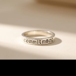 May include: A silver ring with the word "commitment" engraved on the band. The ring is circular and has a simple, elegant design. The text is in a sans-serif font.
