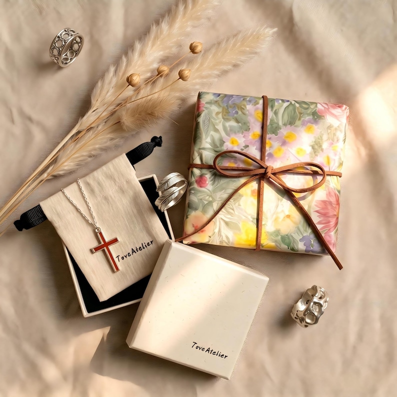 Pu&ograve; includere: Un assortimento di gioielli e regali, tra cui anelli, una collana con pendente a croce e un regalo confezionato. La collana presenta una croce rossa. Il regalo &egrave; avvolto in carta floreale e legato con un nastro marrone. Il marchio "Tove Atelier" &egrave; visibile.