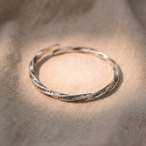 May include: A silver-coloured, twisted metal bracelet with engraved text. The open cuff design features a continuous spiral pattern. The bracelet is displayed on a neutral-toned fabric background, highlighting its reflective surface.