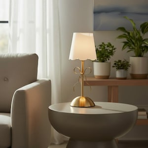 Puede incluir: Una lámpara de mesa con base de metal dorado cepillado que presenta un detalle decorativo de lazo en el vástago y una pantalla blanca con bordes festoneados. La lámpara emite una luz suave y se apoya sobre una base redondeada y lisa.