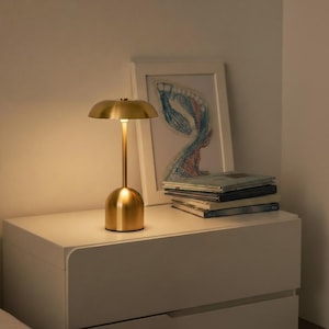 Puede incluir: Una lámpara de mesa dorada a pilas, con una pantalla en forma de hongo y una base cilíndrica. La lámpara está sobre una mesita de noche blanca, junto a un dibujo enmarcado y una pila de libros. La lámpara emite un brillo cálido.