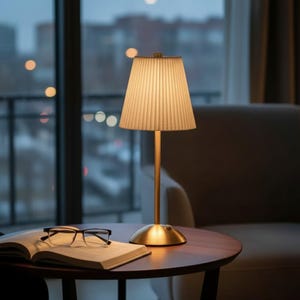 May include: A table lamp with a gold-toned metal base and a tall, slender stem, topped with a cream-colored, pleated fabric lampshade. The lamp emits a warm, soft glow, illuminating a pair of black-framed eyeglasses resting on an open book.