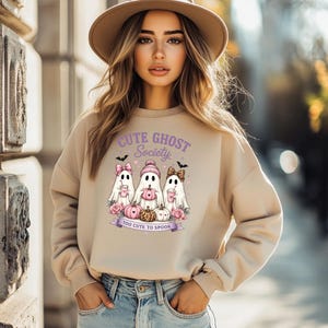 Könnte beinhalten: Ein beigefarbenes Sweatshirt mit Rundhalsausschnitt und einer Grafik von drei Geistern mit rosa Accessoires, Kürbissen sowie dem violetten Text "CUTE GHOST Society" und "TOO CUTE TO SPOOK". Das Design enthält kleine Fledermäuse, Herzen und rosa Rosen.